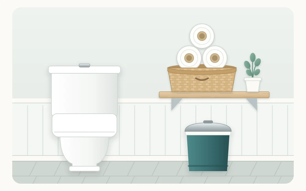 A shelf beside a toilet with three spare rolls of toilet paper stacked in a basket and a small lidded bin below
